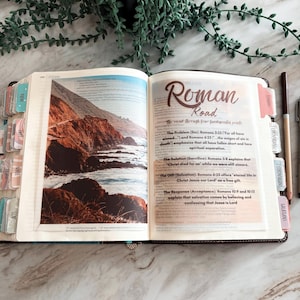 May include: An open Bible featuring a full-page color photograph of a rocky coastline on the left and the text Roman Road on the right page. The book includes colorful index tabs along the edges and a brown pen resting on the right side of the page.