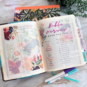 May include: An open Bible with floral patterned vellum pages featuring the text "Bible Verses FOR WHEN YOU FEEL..." followed by a list of emotions in various colors and corresponding scripture references. Two markers lie on the surface below.