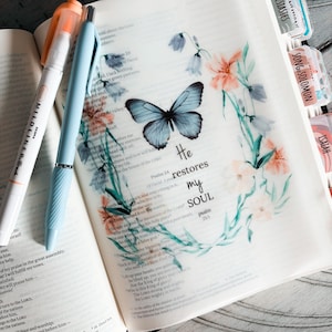 May include: Open Bible with a decorative overlay featuring a blue butterfly and floral accents. The text "He restores my soul" is visible. A light blue pen and an orange highlighter rest on the pages.