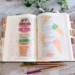 May include: An open Bible with colorful tabs and illustrations of ice cream cones on the pages. The left page features the text "TASTE AND SEE THAT THE LORD IS GOOD" stacked in colorful lettering. Three colored pencils lie horizontally below the book.