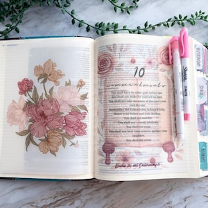 May include: An open book featuring a floral illustration on the left page and the text 10 Commandments on the right page. The pages include colorful index tabs along the edge, and two pink highlighters are placed on the right side of the open book.