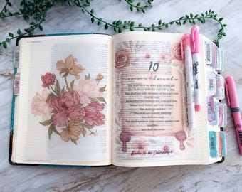 May include: An open book featuring a floral illustration on the left page and the text 10 Commandments on the right page. The pages include colorful index tabs along the edge, and two pink highlighters are placed on the right side of the open book.