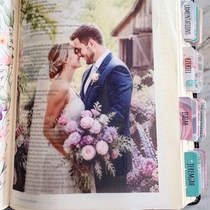 May include: An open book featuring a printed photograph of a couple in wedding attire holding a floral bouquet. The page includes text and several colorful adhesive tabs labeled Lamentations, Ezekiel, Isaiah, and Jeremiah along the edge.