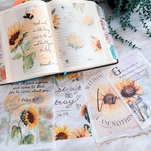 May include: An open Bible with sunflower and bee illustrations, featuring the text Kind words are like honey sweet to the soul and healthy for the body Proverbs 16:24. Below are translucent vellum sheets with sunflower prints and various Bible verses.