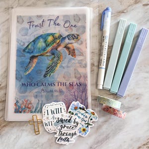 May include: A clear plastic sleeve containing a print of a sea turtle with the text Trust The One WHO CALMS THE SEAS PSALM 46:10, accompanied by four markers, two rolls of patterned washi tape, a gold cross paperclip, and two die-cut stickers.