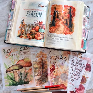May include: An open Bible with autumn-themed illustrations of pumpkins and a forest path, featuring colorful page tabs. Below are translucent vellum sheets with floral and mushroom prints, text reading "rejoice IN THE LORD!", and three colored pencils.