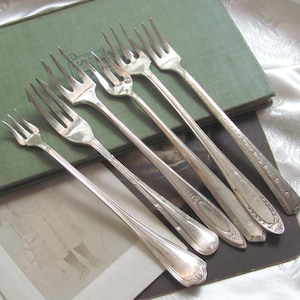 Può includere: Sei forchette d'argento vintage con manici decorati sono disposte in diagonale. Le forchette poggiano su un libro verde e una superficie nera, con un tessuto bianco fantasia sullo sfondo. Un classico set di posate.