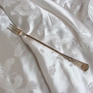 May include: A silver fork with an ornate handle, lying on a white satin fabric with an embossed floral pattern.