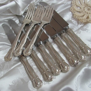 May include: A set of eight silver-plated knives and forks with ornate handles. The silverware is arranged on a white satin fabric background.