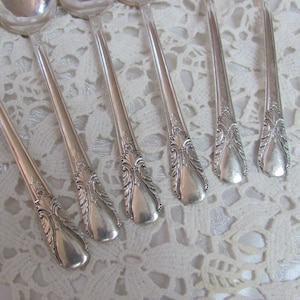 May include: Set of seven silver spoons with ornate handles. The spoons are arranged in a row on a white lace tablecloth.