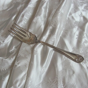 以下が含まれることがあります： 白いサテン生地に置かれた、4つの歯を持つ銀色のフォーク。生地には龍の模様が施されています。フォークの柄には装飾的な花の模様があります。全体的な雰囲気はエレガントでヴィンテージです。