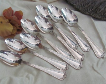 Può includere: Set di otto cucchiai argentati con manici decorati. I cucchiai sono disposti a ventaglio su uno sfondo di tessuto bianco.