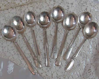 May include: A set of eight silver soup spoons with ornate handles. The spoons are arranged in a fan shape on a white lace tablecloth.