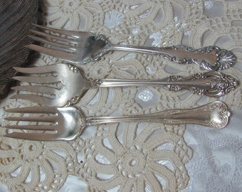 Könnte beinhalten: Drei silberne Gabeln mit verzierten Griffen, auf einer weißen Häkelspitze ausgelegt.