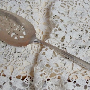 May include: A silver cake server with an ornate, cut-out design. The server is lying on a white lace tablecloth.