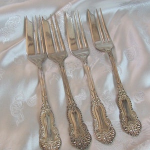 Op de afbeelding: Vier antieke zilveren vorken met sierlijke handvatten en tanden, gerangschikt op een witte, getextureerde ondergrond. De handvatten hebben een krullend ontwerp en een monogram. Deze vorken zijn waarschijnlijk voor formele diners.