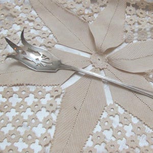 May include: A silver serving fork with a decorative handle rests on a beige floral lace doily. The fork has a pierced design on the tines. The background features a beige floral pattern.