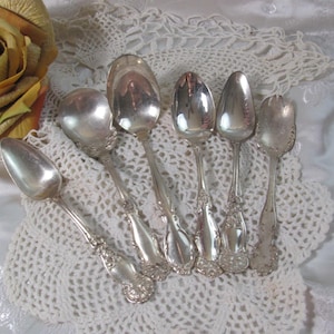 May include: A set of five silver-toned metal spoons with ornate, embossed floral detailing on the handles. The spoons vary in size and shape, featuring polished, reflective bowls and intricate decorative patterns along the stems and handle ends.