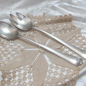 May include: A pair of silver serving utensils, a spoon and a fork, rest on a beige lace doily. The utensils have long, slender handles and are placed on a white satin surface.
