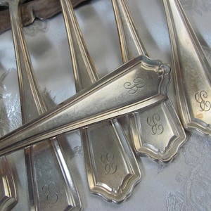 May include: A collection of antique silver-plated spoons and forks. Each piece features a decorative handle with an engraved initial. The silverware is arranged on a patterned, light-colored surface, showcasing the reflective quality of the metal.