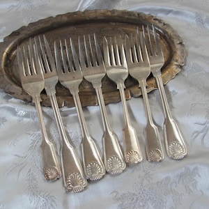 May include: A collection of seven silver forks with ornate handles, arranged on a tarnished, oval metal tray. The forks have a decorative shell design at the base of the handle. The tray sits on a light blue fabric with a subtle pattern.