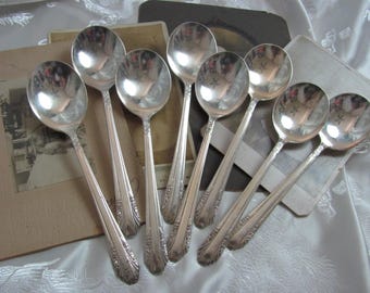 May include: Set of eight silver-plated soup spoons with ornate handles. The spoons are arranged in a fan shape on a brown surface.