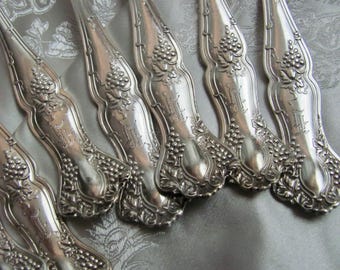 May include: Close-up of several silver-colored forks. The forks have ornate handles with a grape and leaf design. The handles are curved and have a detailed, embossed pattern. The forks are arranged on a patterned, light gray surface.