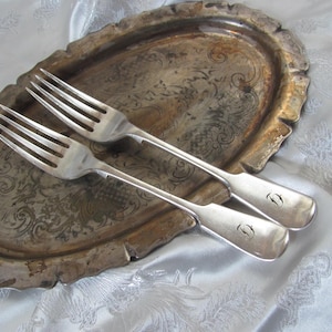 May include: Two silver forks rest on a tarnished, oval silver tray. The tray has an ornate, embossed design and a scalloped edge. The forks have a simple design with four tines and a curved handle. The background is a white, patterned fabric.
