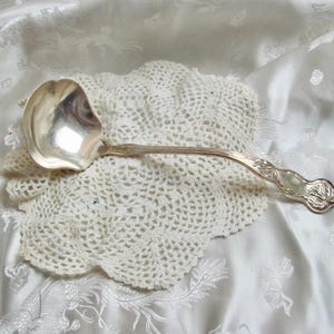 May include: A silver ladle with an ornate handle rests on a cream-coloured, crocheted doily. The ladle's bowl reflects light, and the handle features a floral design. The background is a white, textured fabric.