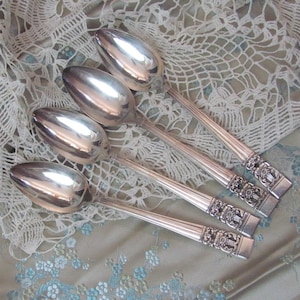 Pode incluir: Cinco colheres de prata com detalhes ornamentais na alça. As colheres têm uma superfície polida e reflexiva e são dispostas sobre um tecido estampado com uma sobreposição de renda. As alças apresentam um design floral e geométrico.