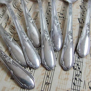 May include: A collection of antique silver spoons with ornate floral and leaf designs. The spoons are arranged on a vintage sheet music background, showcasing their elegant, vintage aesthetic. The spoons are made of silver.