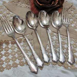 May include: A set of six antique silver-plated spoons and forks. The silverware has ornate floral detailing on the handles. The spoons and forks are arranged on a cream-colored lace doily.