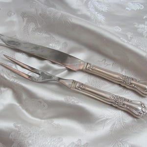 Puede incluir: Juego de trinchar plateado, que incluye un cuchillo y un tenedor, con detalles florales ornamentados en los mangos. El juego se muestra sobre una tela blanca estampada, ideal para una cena formal.