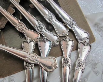 Puede incluir: Primer plano de cubiertos de plata antiguos, que incluyen cucharas y tenedores. La plata presenta diseños florales y de volutas ornamentados, con algunas piezas que muestran iniciales grabadas. Las piezas están dispuestas sobre un fondo vintage.