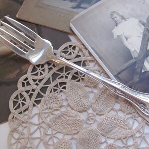 May include: A silver serving fork with ornate detailing rests on a beige lace doily. Vintage photographs of a baby and a woman are in the background. The fork has four tines and a decorative handle, showcasing a classic design.