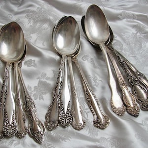 May include: A set of silver spoons with ornate handles. The spoons are arranged in a fan shape on a white fabric background.