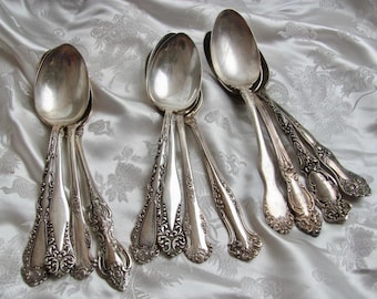 May include: A set of silver spoons with ornate handles. The spoons are arranged in a fan shape on a white fabric background.