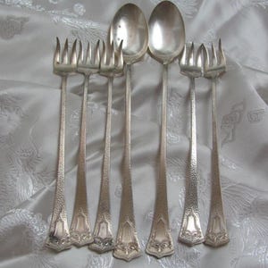 May include: Silverware set featuring two spoons and four forks. The spoons have oval bowls and long, slender handles. The forks have four tines and matching handles. The handles have a textured pattern and decorative detailing at the base.