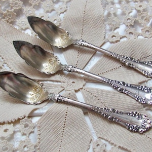 Pode incluir: Três colheres de servir de prata com cabos ornamentados e um design decorativo. As colheres estão dispostas sobre uma superfície de crochê branca.