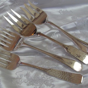 May include: Four silver serving forks with hammered handles. The forks have different numbers of tines and are arranged on a white fabric background.