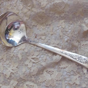 May include: A silver ladle with a floral design on the handle. The ladle is on a white lace background.