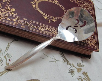 May include: Silver serving spoon with an ornate design and openwork pattern. The spoon is resting on a closed book with a gold-embossed cover.