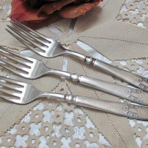 May include: Three antique silver forks with ornate handles are arranged on a beige lace tablecloth. The forks have tines and a decorative design at the end of the handle. A red flower is in the background.
