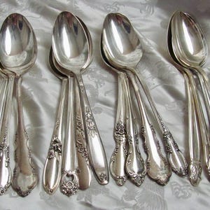 May include: A set of eight silver spoons with ornate handles. The spoons are arranged in a row on a white fabric background.