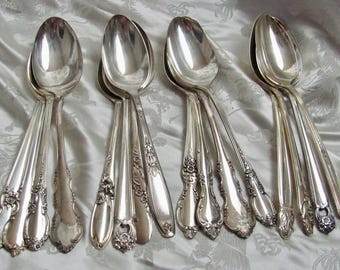 Puede incluir: Un juego de ocho cucharas de plata con mangos adornados. Las cucharas están dispuestas en fila sobre un fondo de tela blanca.
