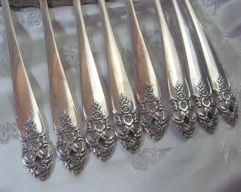 May include: A set of seven silver-toned metal spoons arranged in a parallel row. Each spoon handle features a detailed embossed floral pattern with roses and leaves at the base, transitioning into a smooth, plain stem toward the bowl of the spoon.