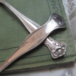 May include: Two silver spoons, one with an ornate floral design and the other with the word "Herbrat" engraved on the handle.