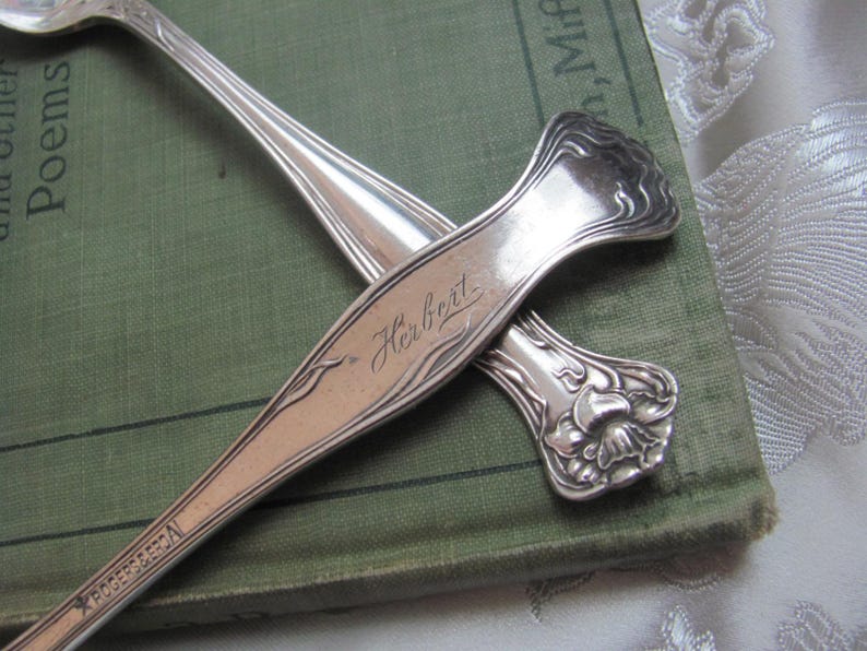 May include: Two silver spoons, one with an ornate floral design and the other with the word "Herbrat" engraved on the handle.