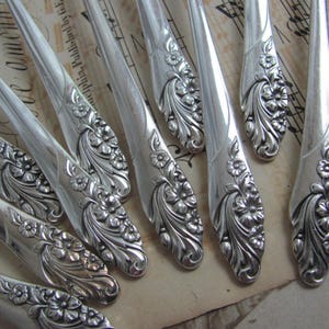 May include: Close-up of several silver-colored forks with floral designs on the handles. The forks are arranged on a vintage paper background with musical notes and text. The floral pattern includes detailed flowers and leaves.