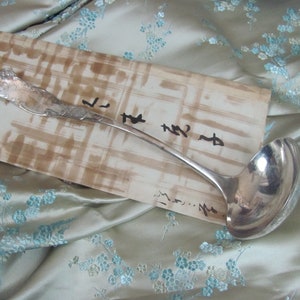 May include: A silver ladle with an ornate handle on a brown and white patterned surface. The surface has handwritten text on it.